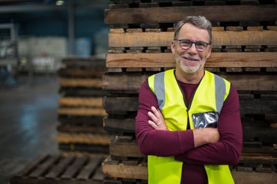 Smiling older industrial worker leaning against a stack of pallets